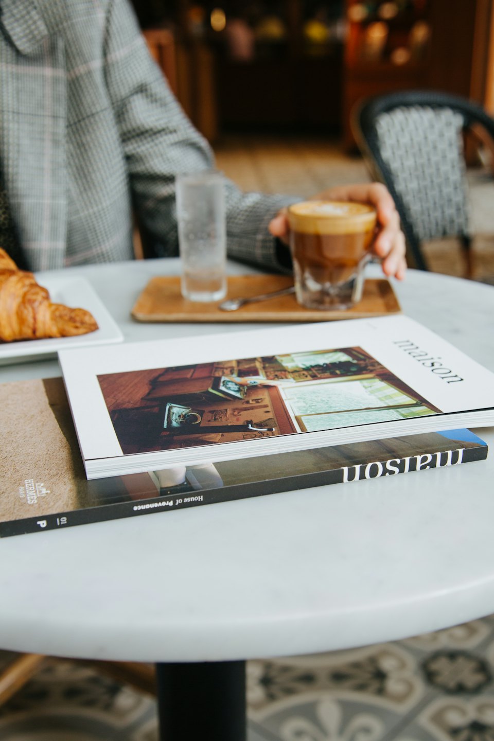 Person in plaid jacket reads magazines at marble table with coffee.