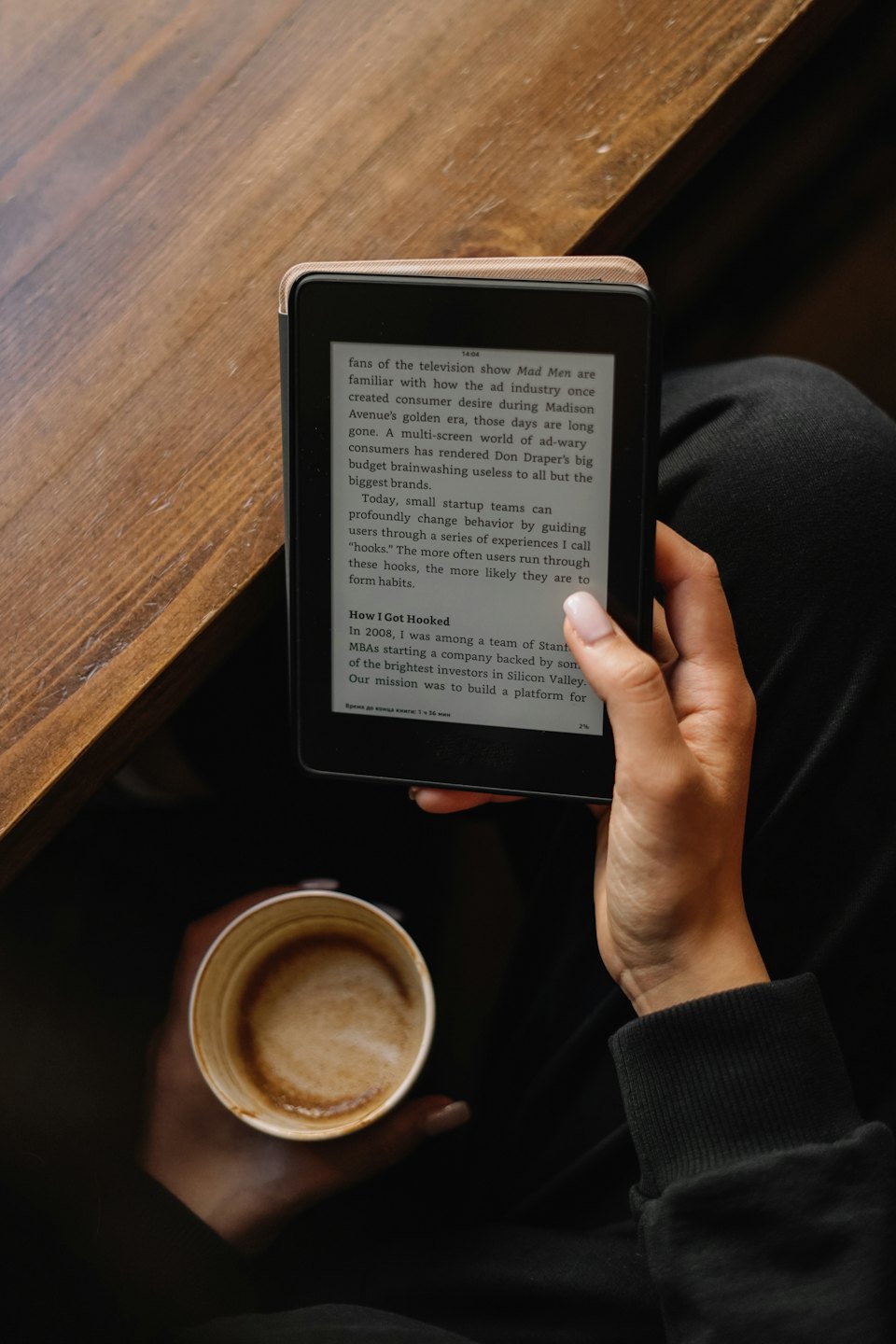 Person holding an e-reader and a coffee cup on a wooden table.