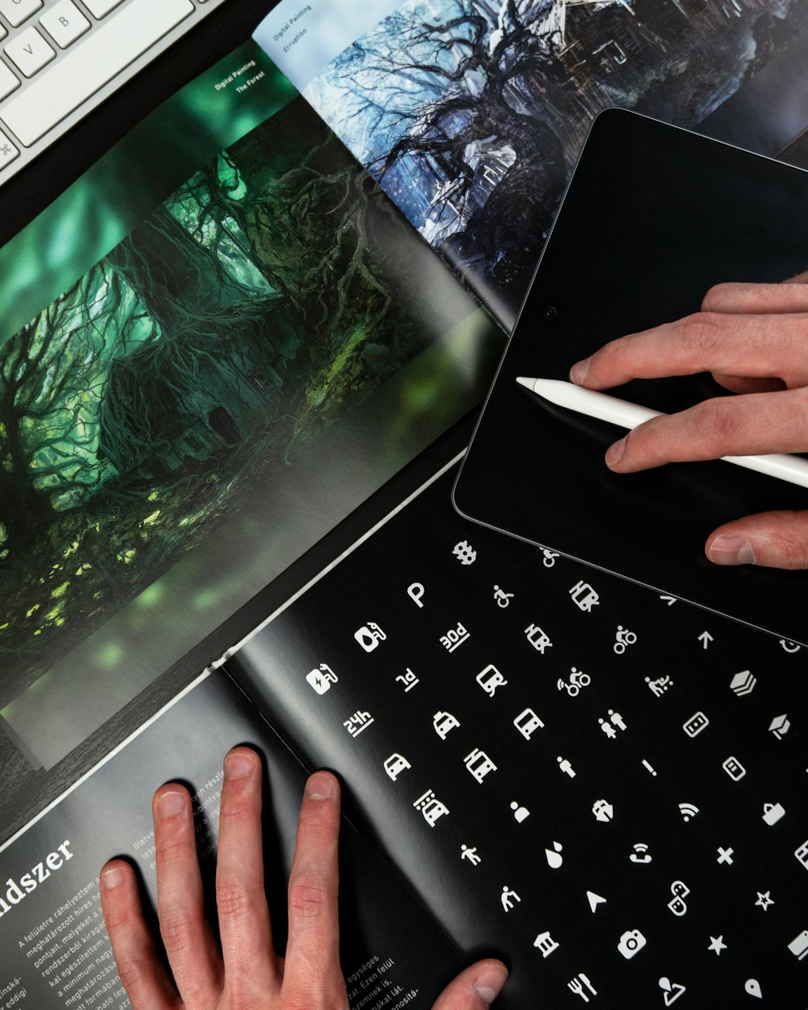 Hands using a stylus on a tablet next to a magazine and keyboard.